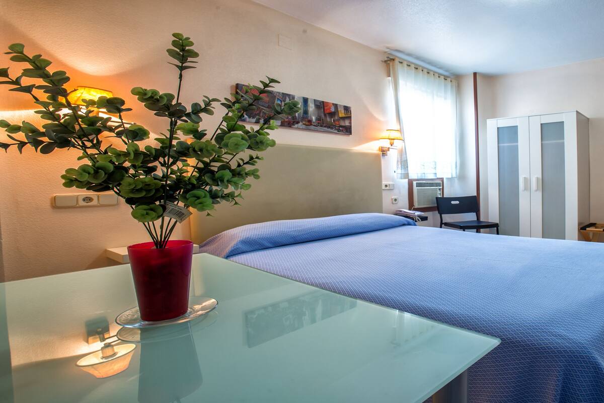 A hotel room with a bed, a desk with a red vase, a painting on the wall, and a wardrobe.