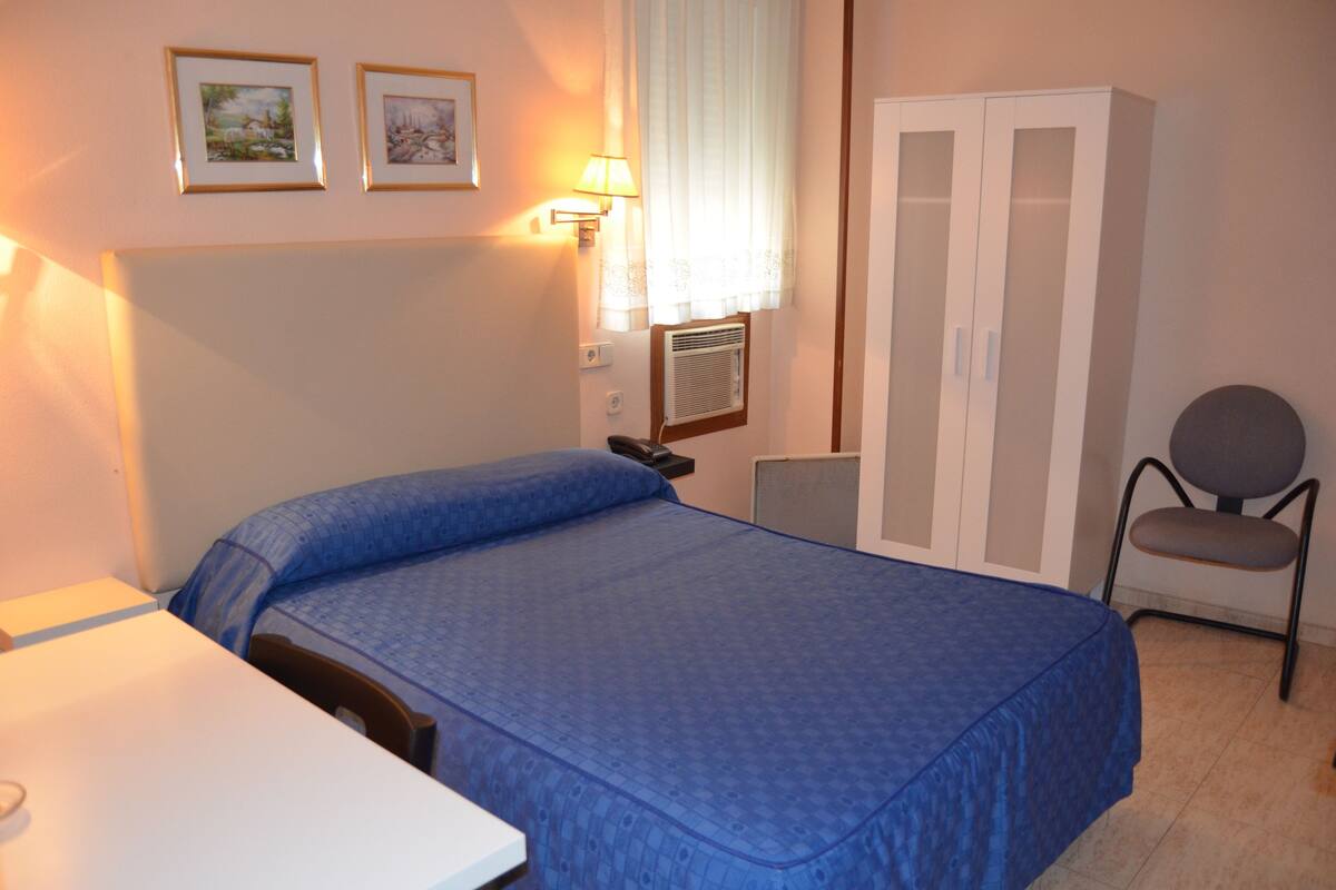 A bedroom with a bed, a desk, a chair, a wardrobe, and two wall-mounted lamps.