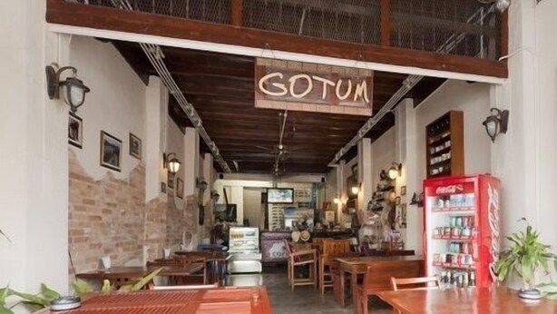 A cozy restaurant interior with wooden tables and chairs, a Coca-Cola vending machine, and a sign reading "GOTUM". - Gotum Hostel & Restaurant (Phuket)