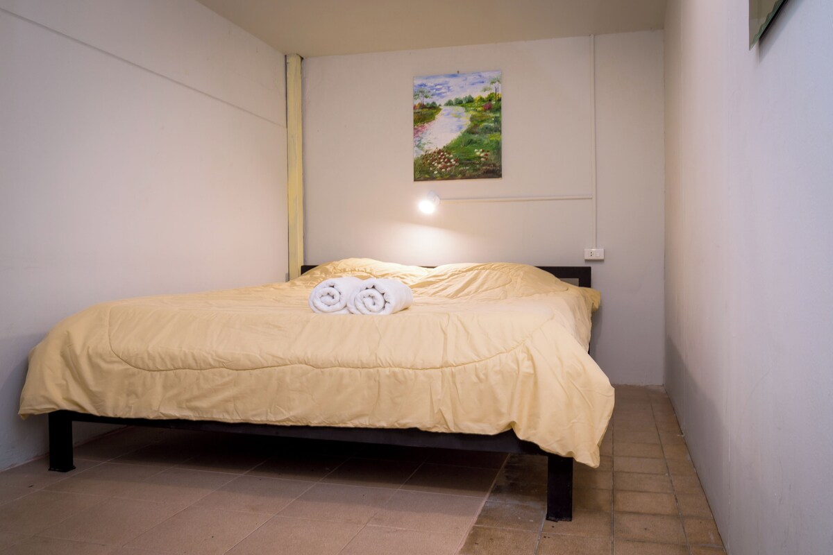 A single bed with a yellow bedspread and two rolled towels placed on it.