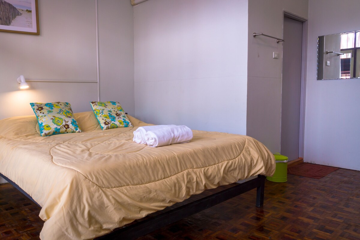 A bed with a beige bedspread and two floral pillows, a rolled white towel, and a green trash bin.