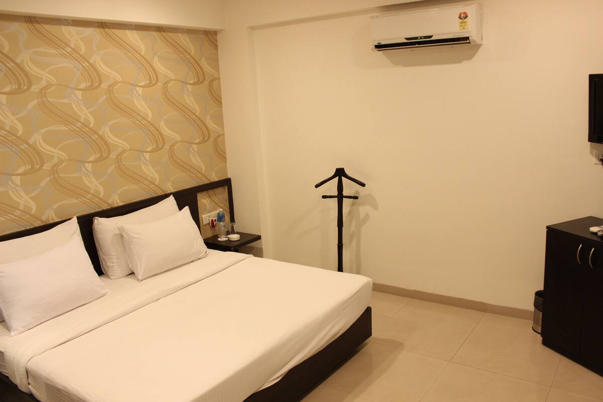 A hotel room with a bed, a nightstand, an air conditioning unit, a black cabinet, and a patterned wallpaper.