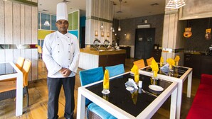 L’intérieur d’un restaurant moderne présente un chef debout près d’une table dressée pour quatre personnes, avec des chaises bleues, une table noire et des serviettes jaunes.