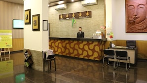 Un espace d’accueil d’hôtel comprenant un comptoir de réception avec un réceptionniste, un écran numérique, des œuvres d’art encadrées et une statue de Bouddha.