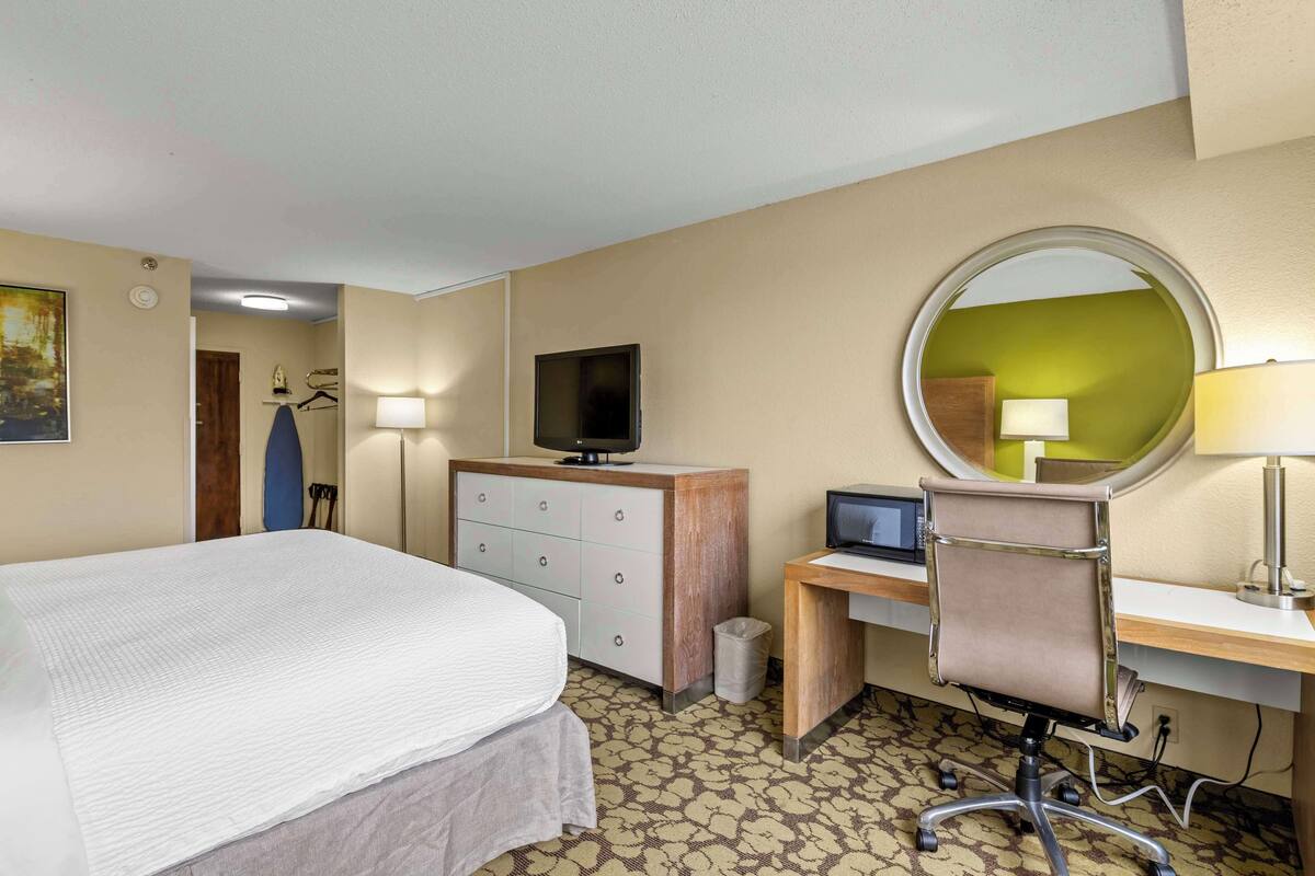 A hotel room with a bed, a desk, a chair, a television, a mirror, and a lamp.