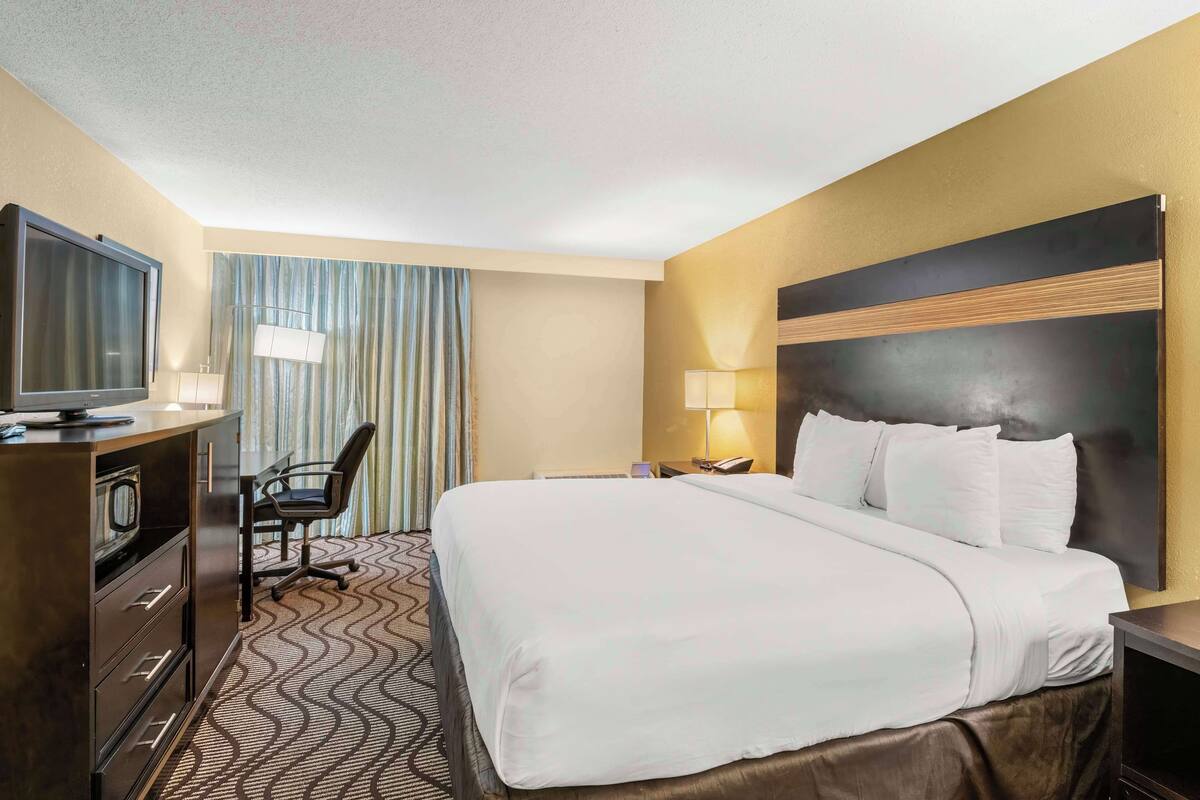A hotel room with a large bed, a desk with a television, a chair, a desk lamp, and a nightstand with a phone.