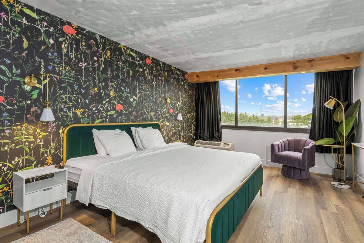 A bedroom with a floral wallpaper, a green upholstered headboard, a white bedspread, a purple armchair, and a large window with a view.