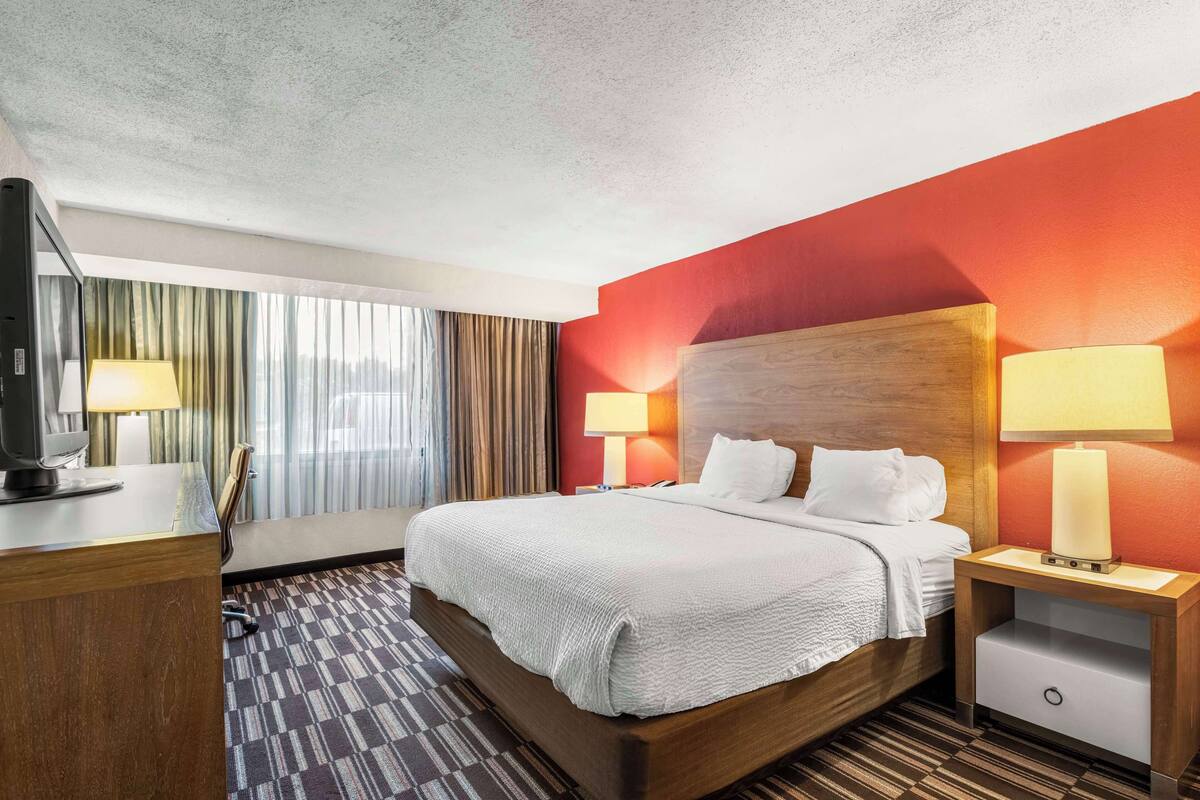 A hotel room with a large bed, two bedside lamps, a wooden headboard, a desk with a computer, and a view of the outdoors through curtains.