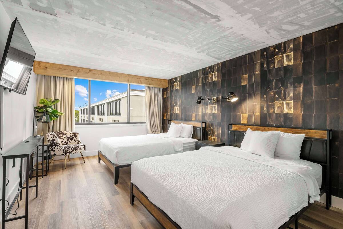 A hotel room with two beds, a desk, a chair, and a large window offering a city view.
