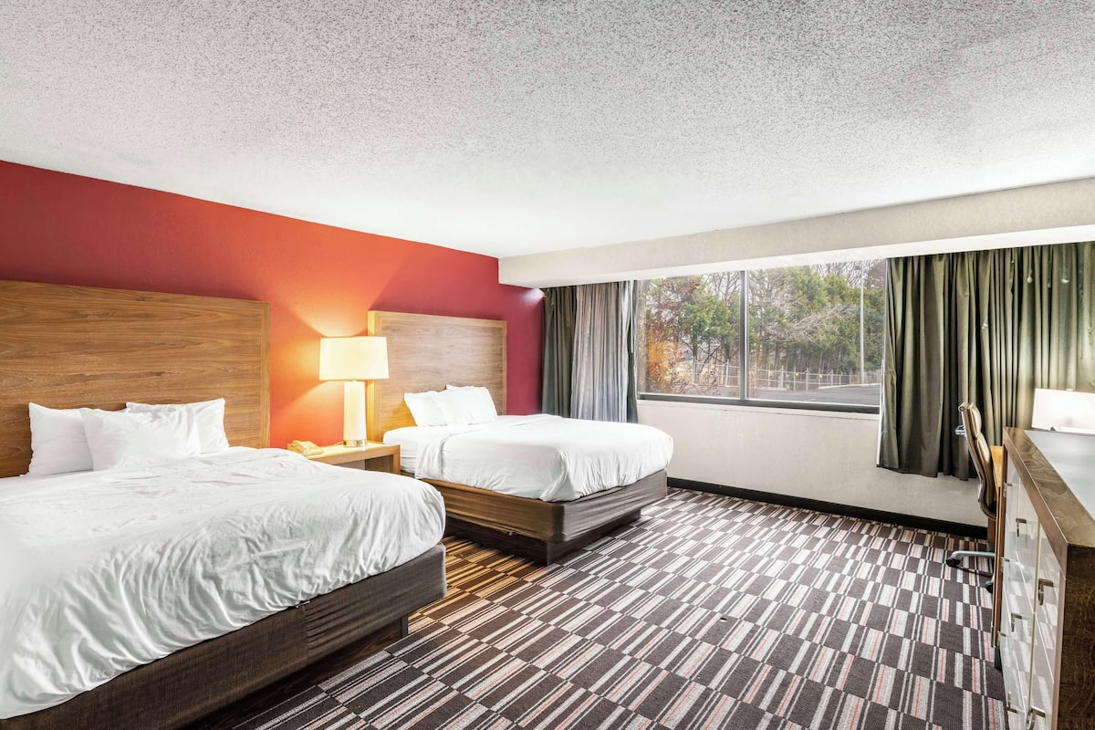 A hotel room with two beds, a wooden headboard, a nightstand with a lamp, a desk with a chair, and a view of trees through the window.