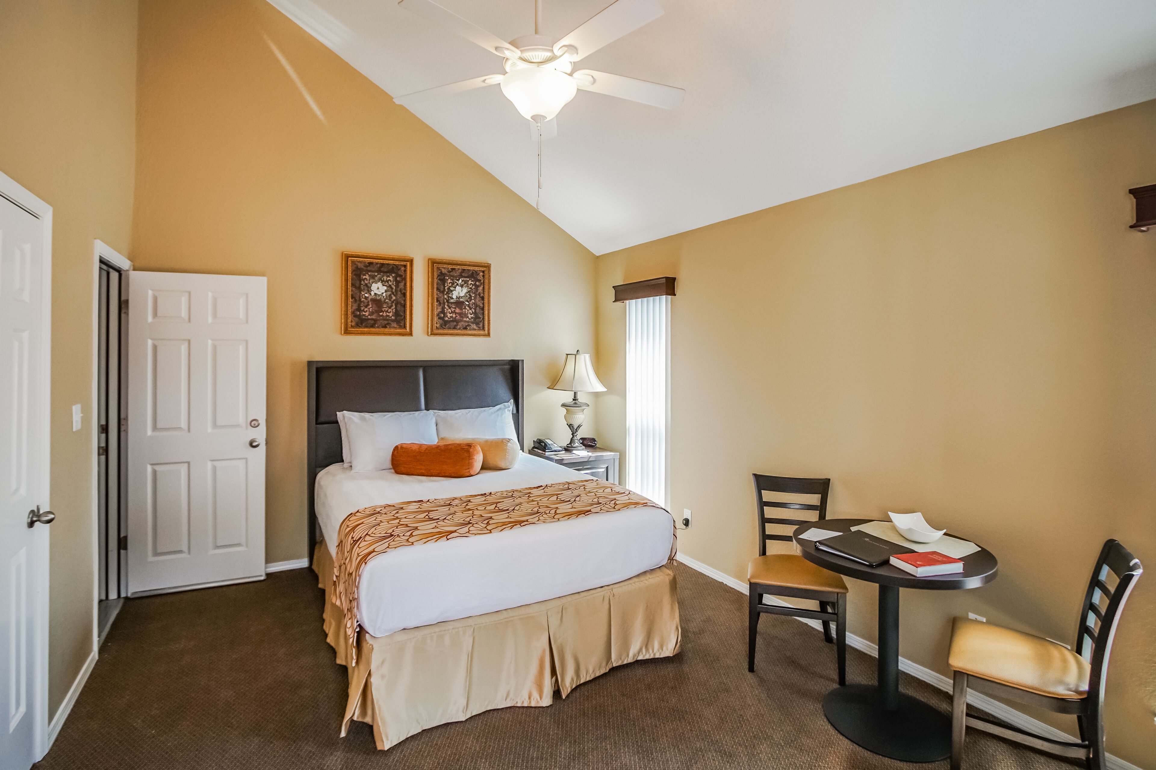 A hotel room with a bed, a desk, a chair, a ceiling fan, and two framed pictures on the wall.