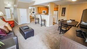 A spacious living room with a dark leather sofa, a coffee table, and a dining area with a wooden table and chairs. A kitchen with wooden cabinets and a refrigerator is visible in the background. - The Townhouses (Branson)