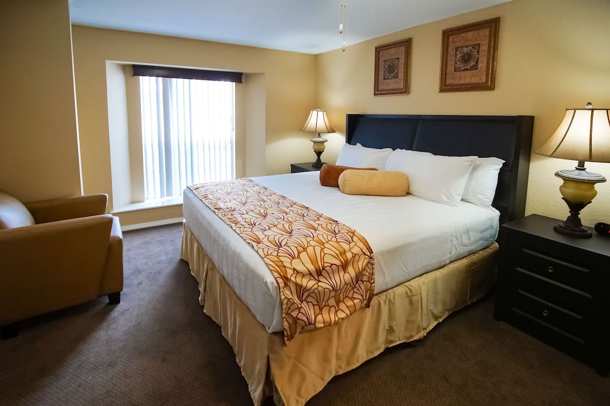 Une chambre d’hôtel avec un grand lit, deux tables de chevet, une chaise et une fenêtre avec des stores.