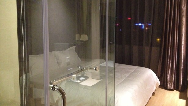 Una habitación de hotel moderna con un separador de vidrio, una cama con ropa blanca, una mesita de noche y una lámpara.