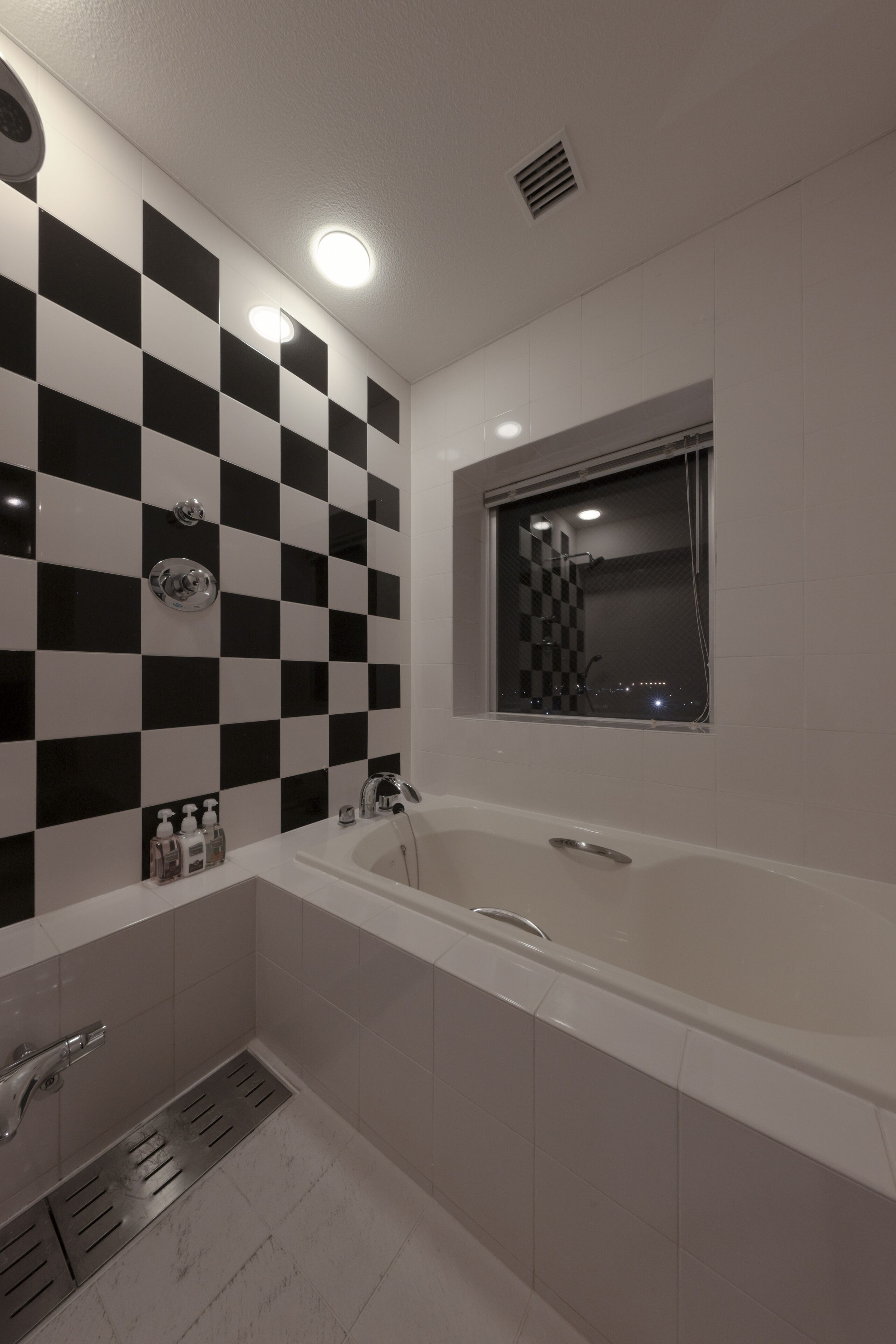 A modern bathroom with a checkered black and white tile wall, a large bathtub, and a window with a view of the city at night.
