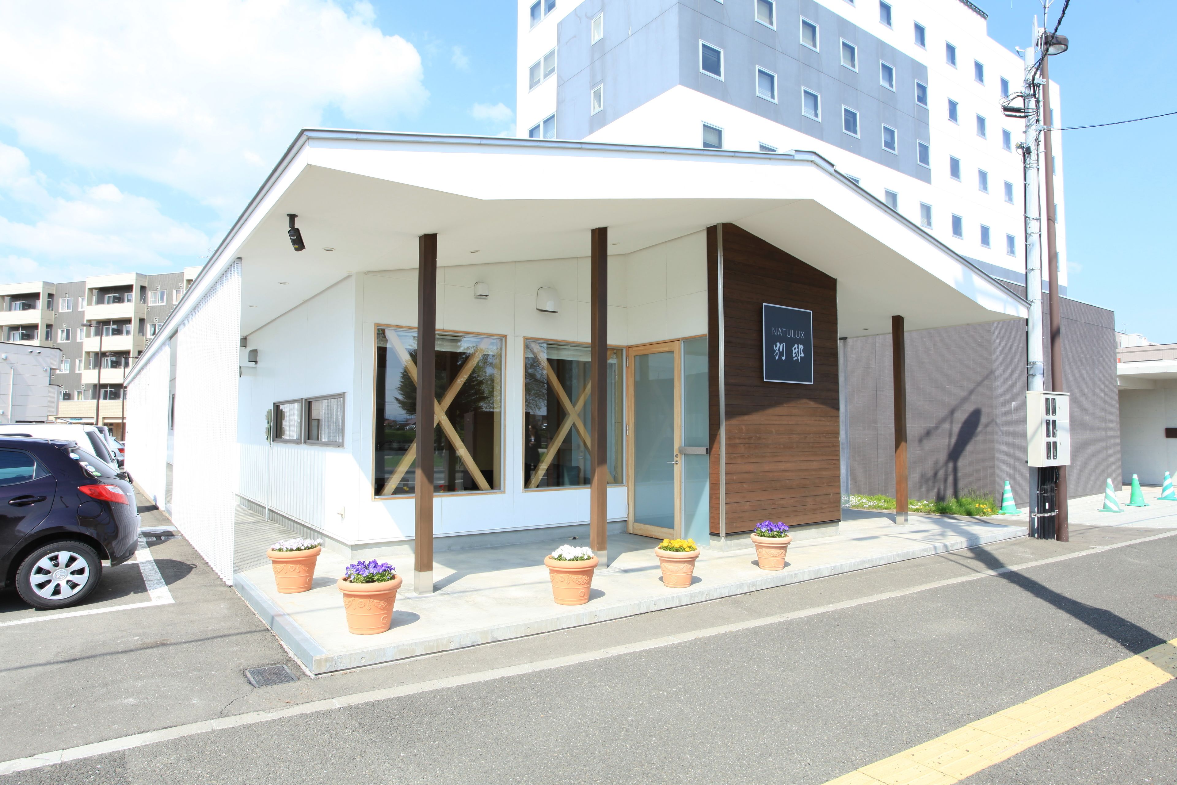 A modern building with a white facade, large windows, and a wooden door. There are potted plants on the sidewalk.