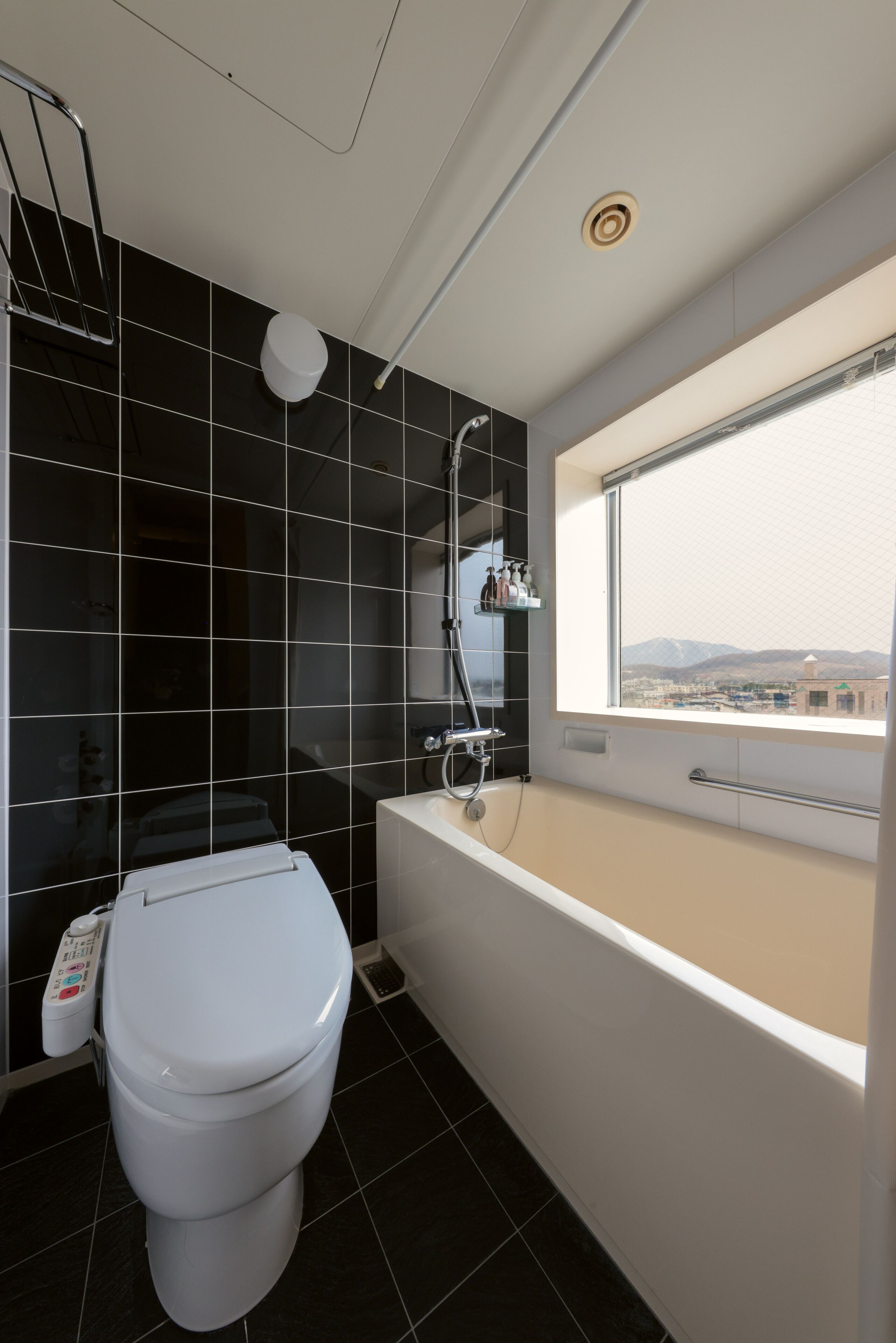 A modern bathroom with a toilet, bathtub, and a window offering a city view.