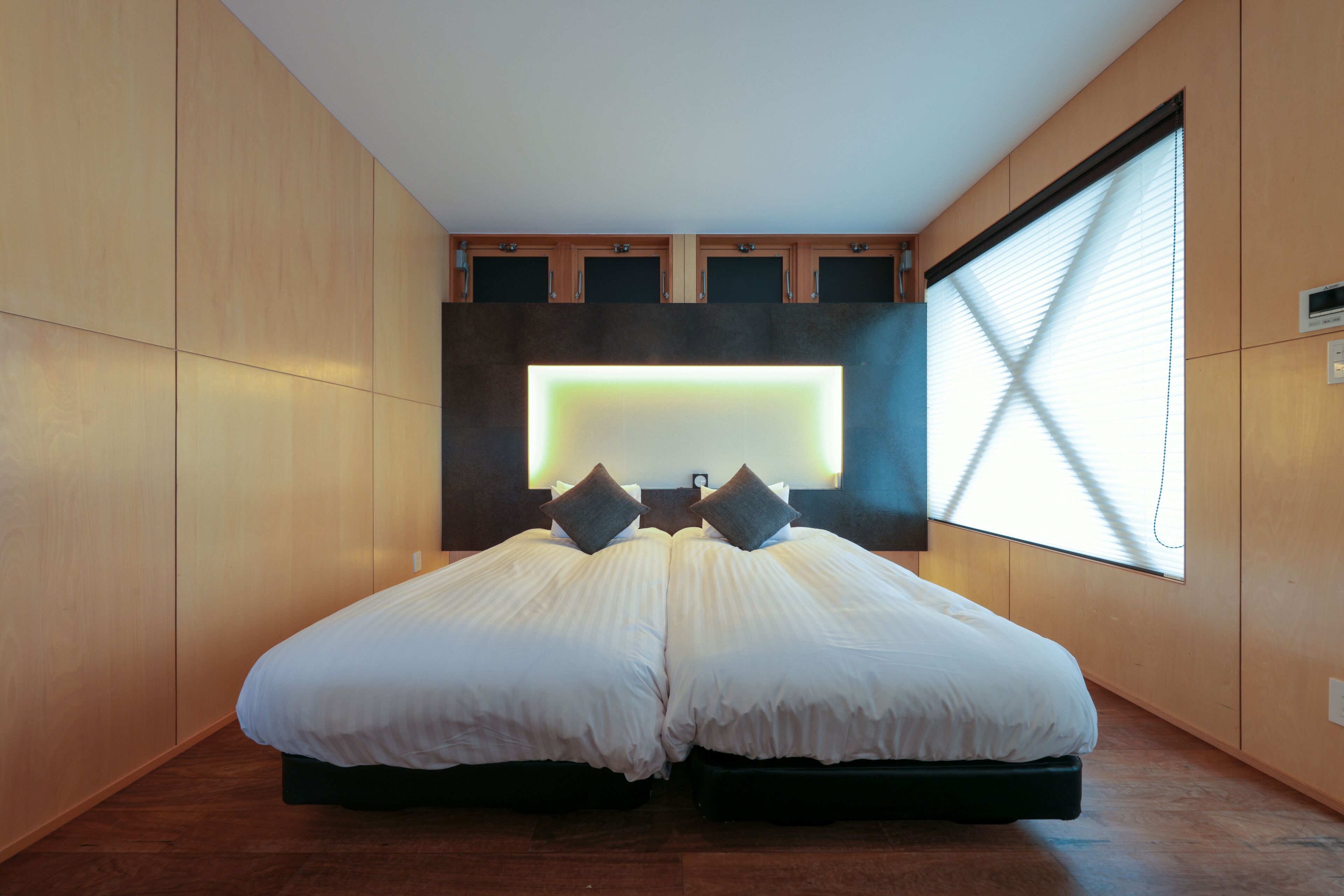A modern hotel room with a large bed, built-in cabinets, and a large window with blinds.