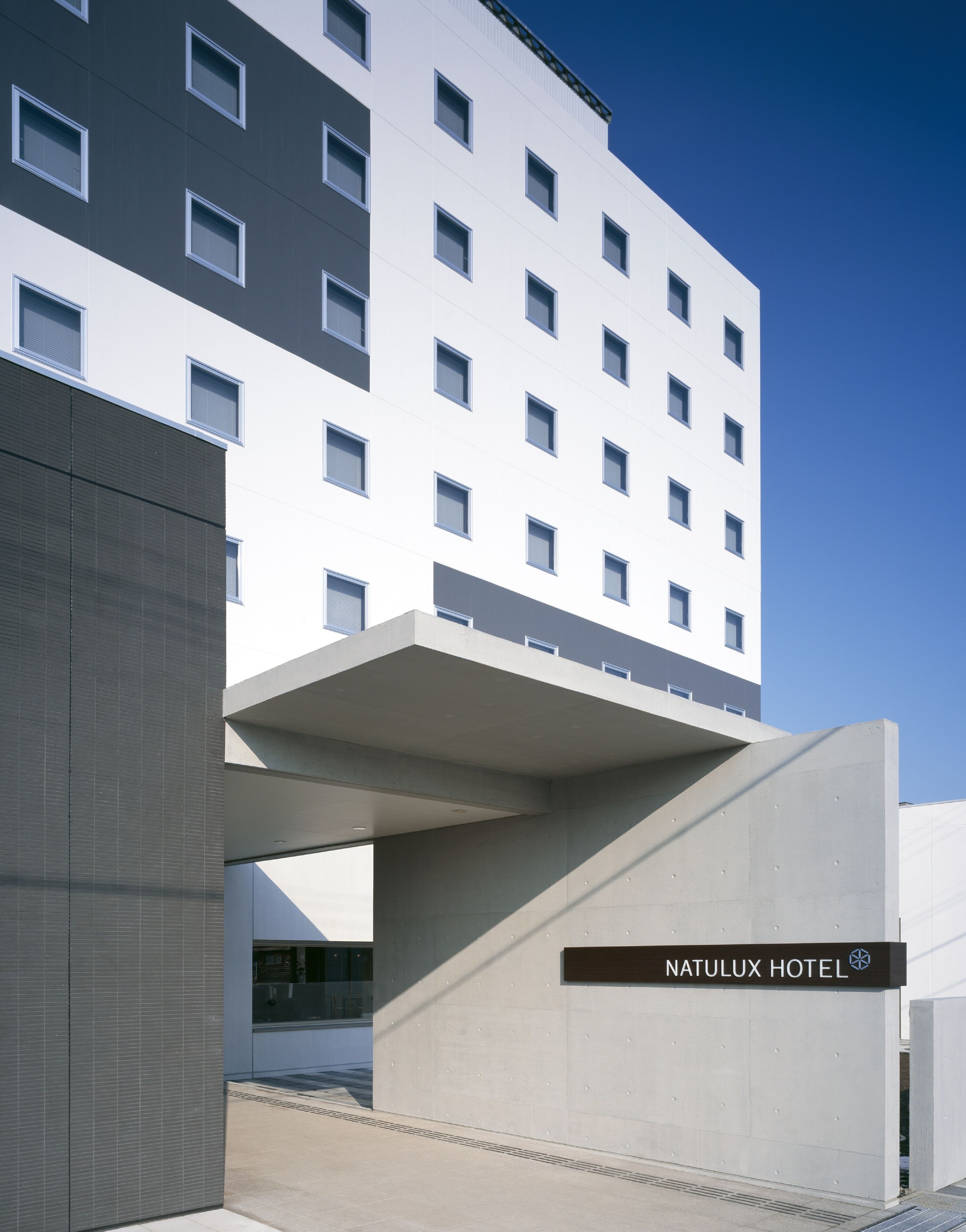 Un moderno edificio de hotel con fachada plana, múltiples ventanas y una entrada destacada con un letrero que dice “NATULUX HOTEL”.
