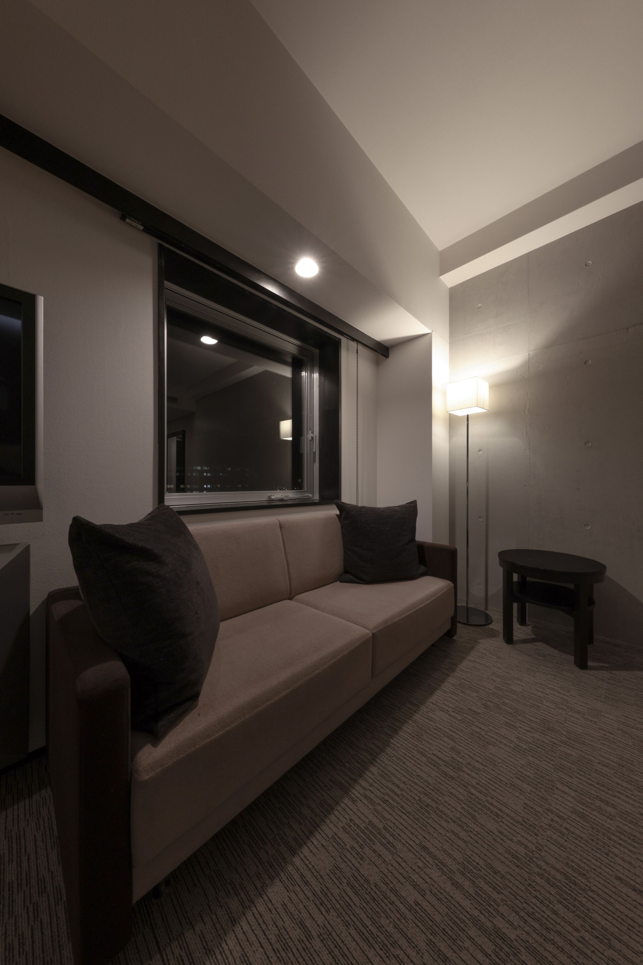 Una sala de estar moderna con un sofá beige, dos cojines oscuros, una lámpara de pie y una mesita.