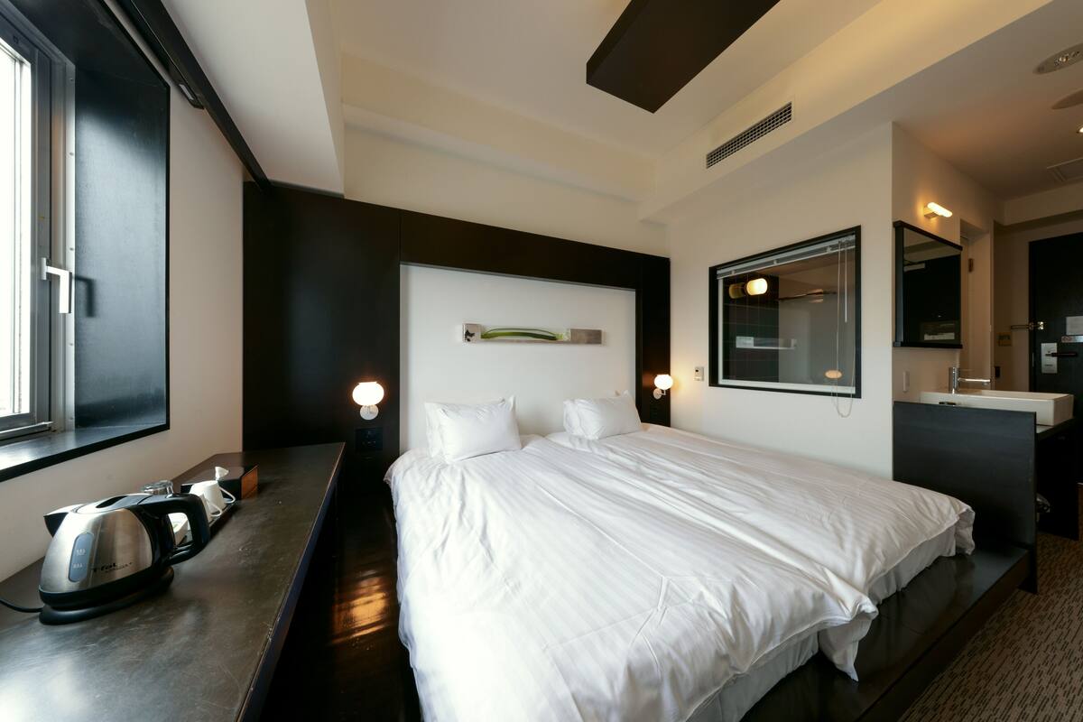 A modern hotel room with a large bed, a small kitchenette, a mirror, and a window.
