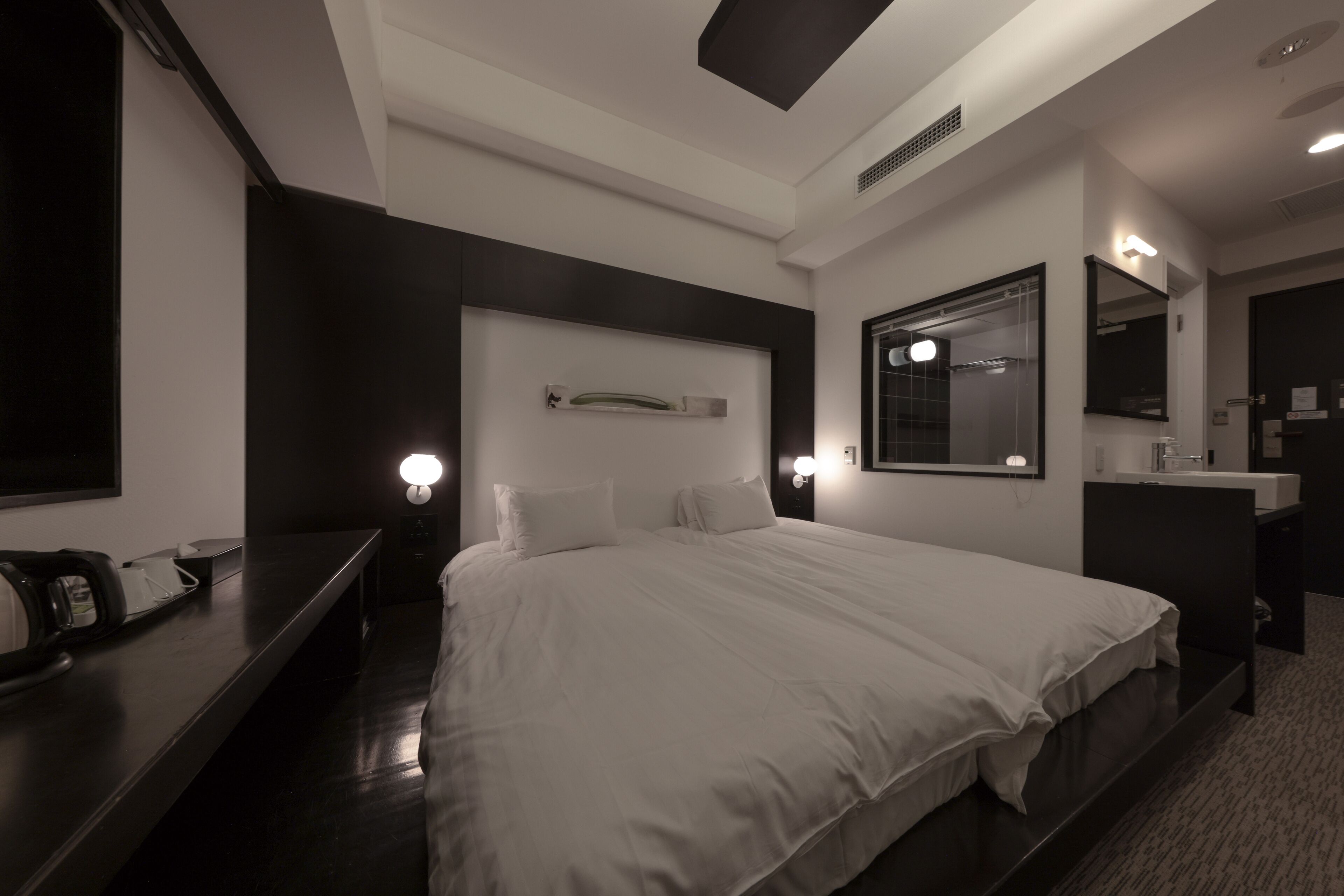 A modern hotel room with a large bed, bedside tables, a mirror, and a small shelf with decorative items.