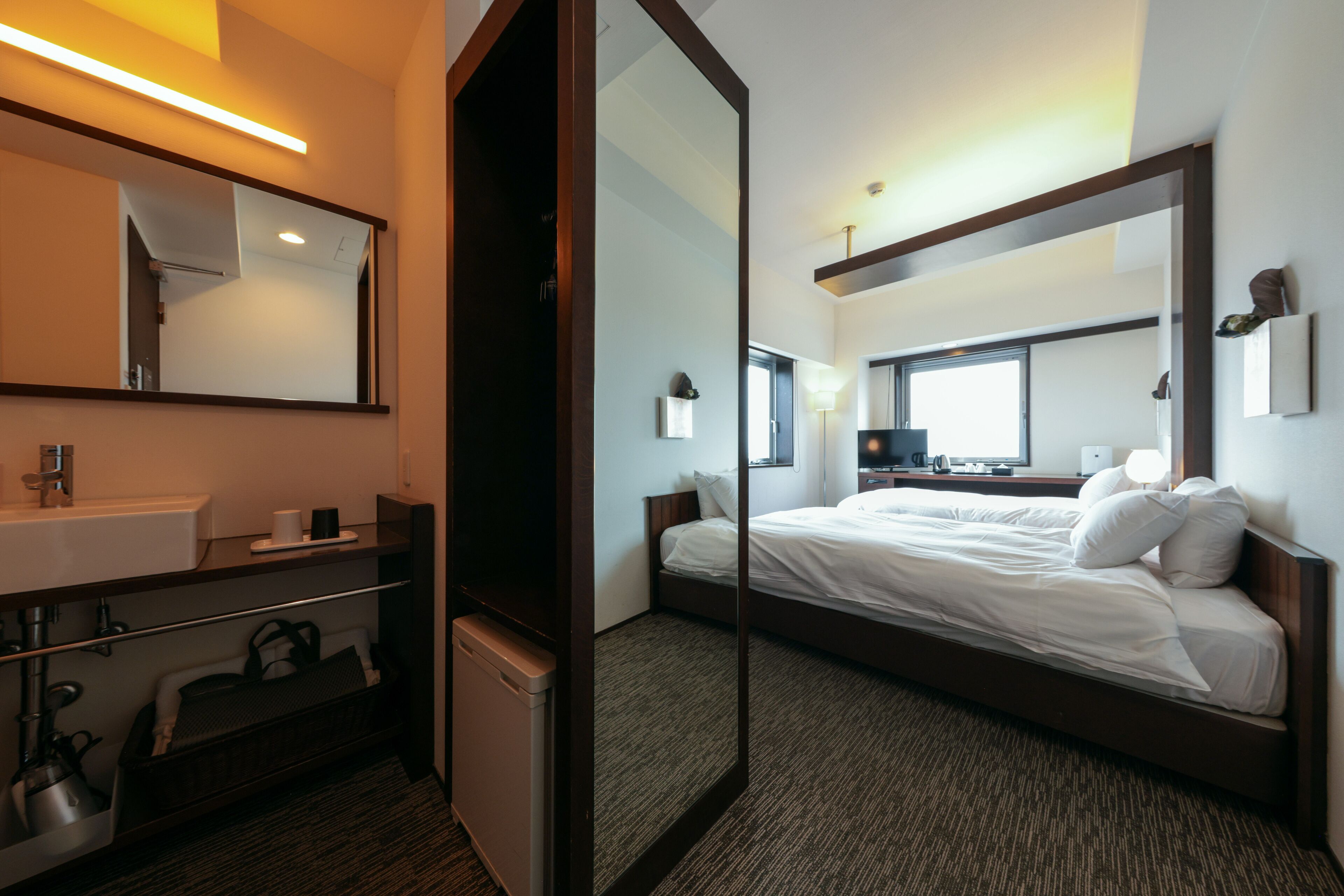 A hotel room with a large bed, a bedside table, a mirror, a sink, and a window.
