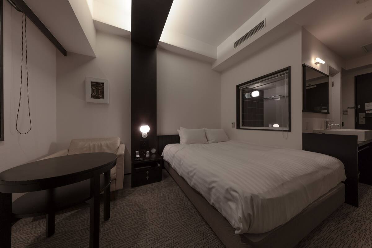 A hotel room with a bed, a desk, a chair, a nightstand, a wall-mounted mirror, and a picture on the wall.