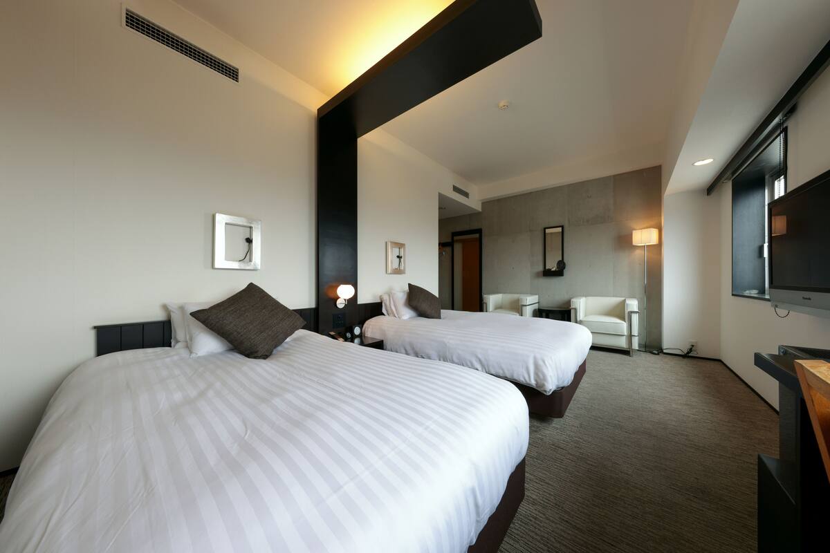 A hotel room with two beds, a desk, a chair, a TV, and a lamp.