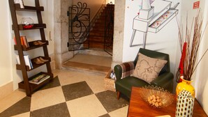 A checkered floor, a wooden table with a vase and a tray, a green sofa with a cushion, a wooden ladder shelf, and a wall with a colorful mural. - Hall Chiado (Lisbon)