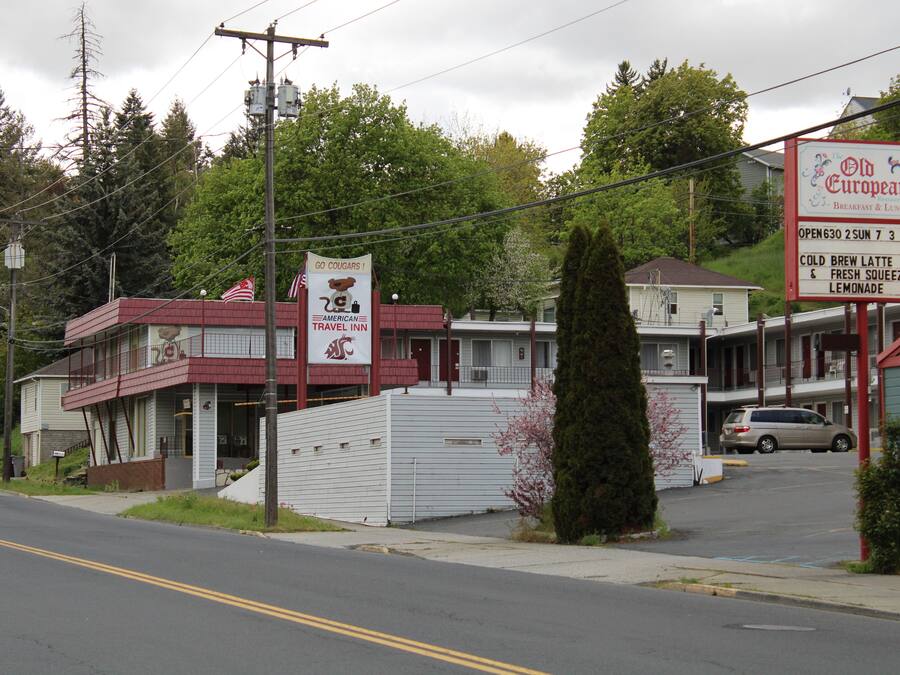 Un motel con fachada roja y blanca, un letrero que anuncia 'Old European Breakfast & Lunch', y un estacionamiento con un coche.