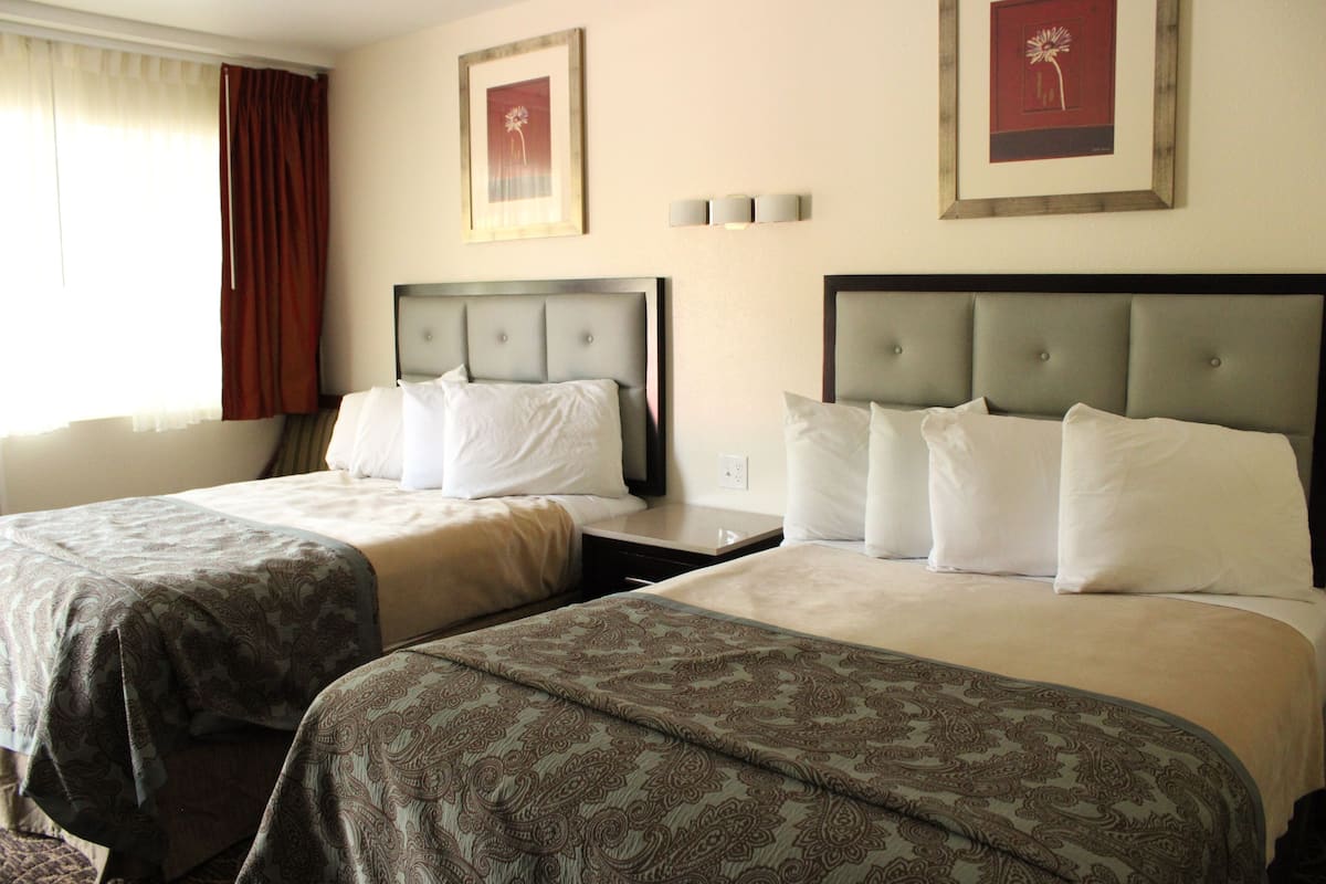 A hotel room with two beds, each with a headboard and pillows. There are framed pictures on the wall and a window with curtains.