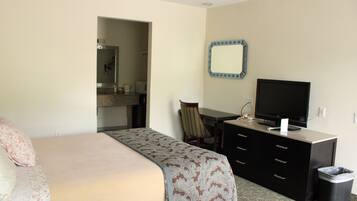 A hotel room with a bed, a desk with a television, a chair, a dresser, and a trash can.