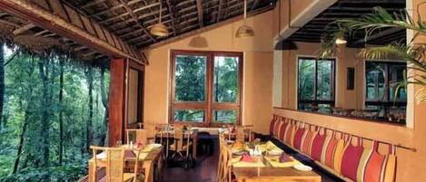 An indoor dining area with wooden tables and chairs, large windows, and a view of the forest.