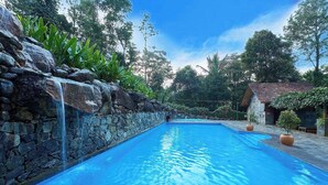 Una piscina con pared de piedra y una cascada a un lado, rodeada de árboles y un edificio con techo de tejas.