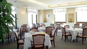 Un comedor con varias mesas y sillas, ventanas amplias con cortinas blancas e iluminación empotrada en el techo.