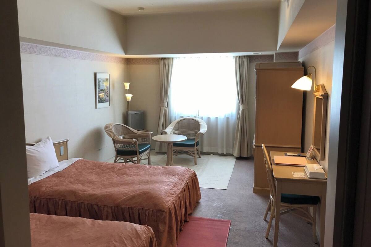 A hotel room with two beds, a desk, a chair, a small table, a lamp, and a window with curtains.
