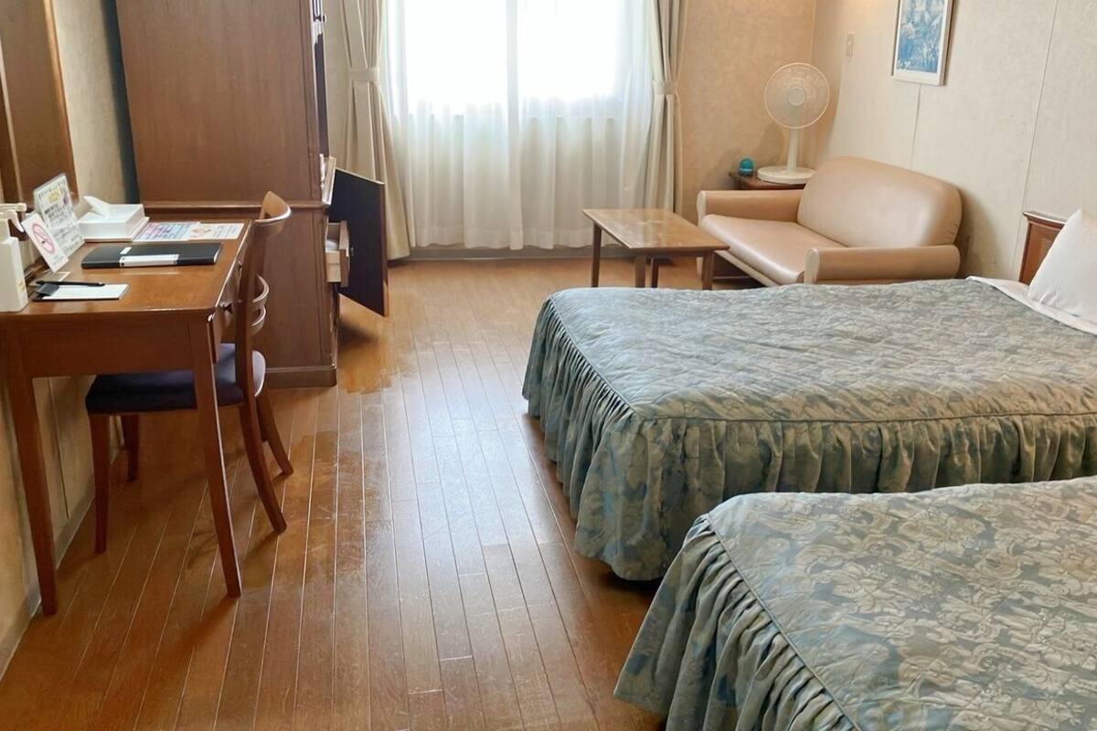 A hotel room with two beds, a desk with a chair, a small table, a sofa, and a fan.