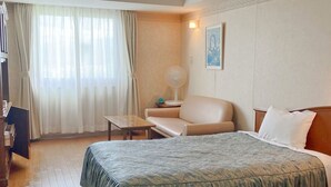 Una habitación de hotel con cama, una silla, una mesita, un ventilador y una ventana con cortinas.