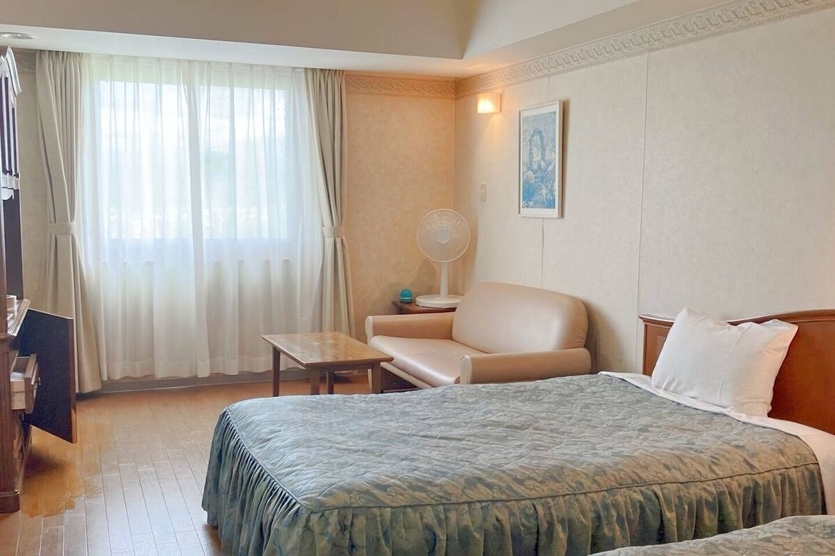 A hotel room with a bed, a chair, a small table, a fan, and a window with curtains.