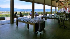 Un espacio de exhibición interior con mesas mostrando artículos, amplias ventanas que ofrecen vistas al exterior y un techo alto con vigas visibles.