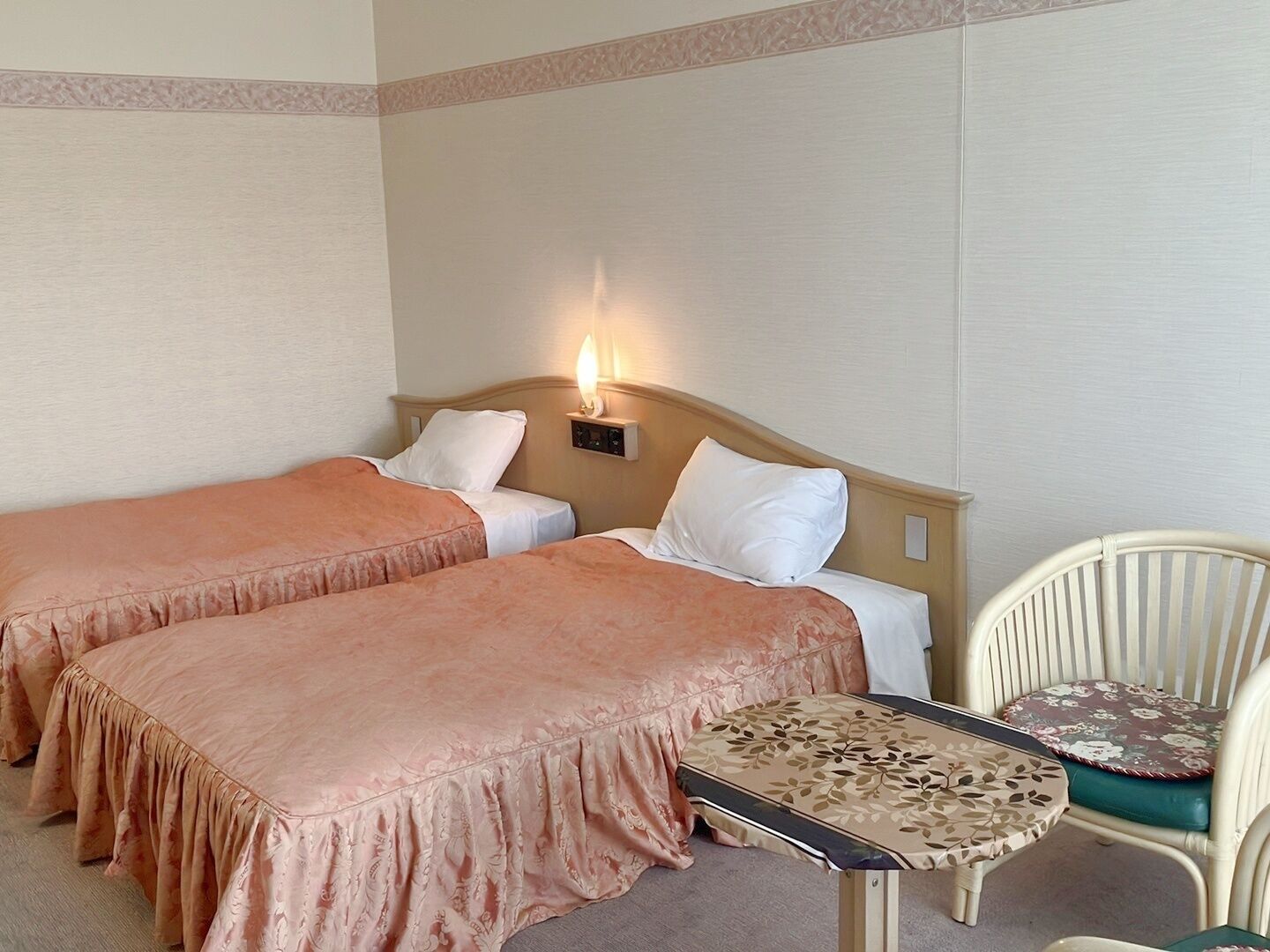 A hotel room with two single beds, a small table, a chair, and a nightstand with a lamp.