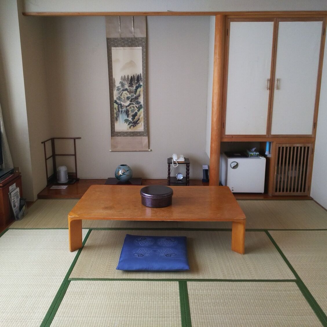 A traditional Japanese room with tatami flooring, a low wooden table, a blue cushion, a small shelf with a vase, a painting, and a wardrobe.