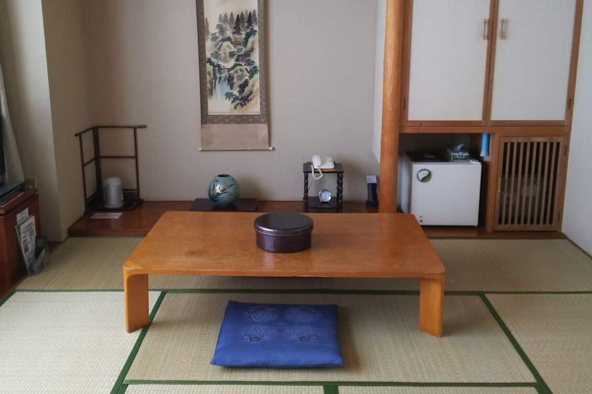 A traditional Japanese room with tatami flooring, a low wooden table, a blue cushion, a small shelf with a vase, a painting, and a wardrobe.