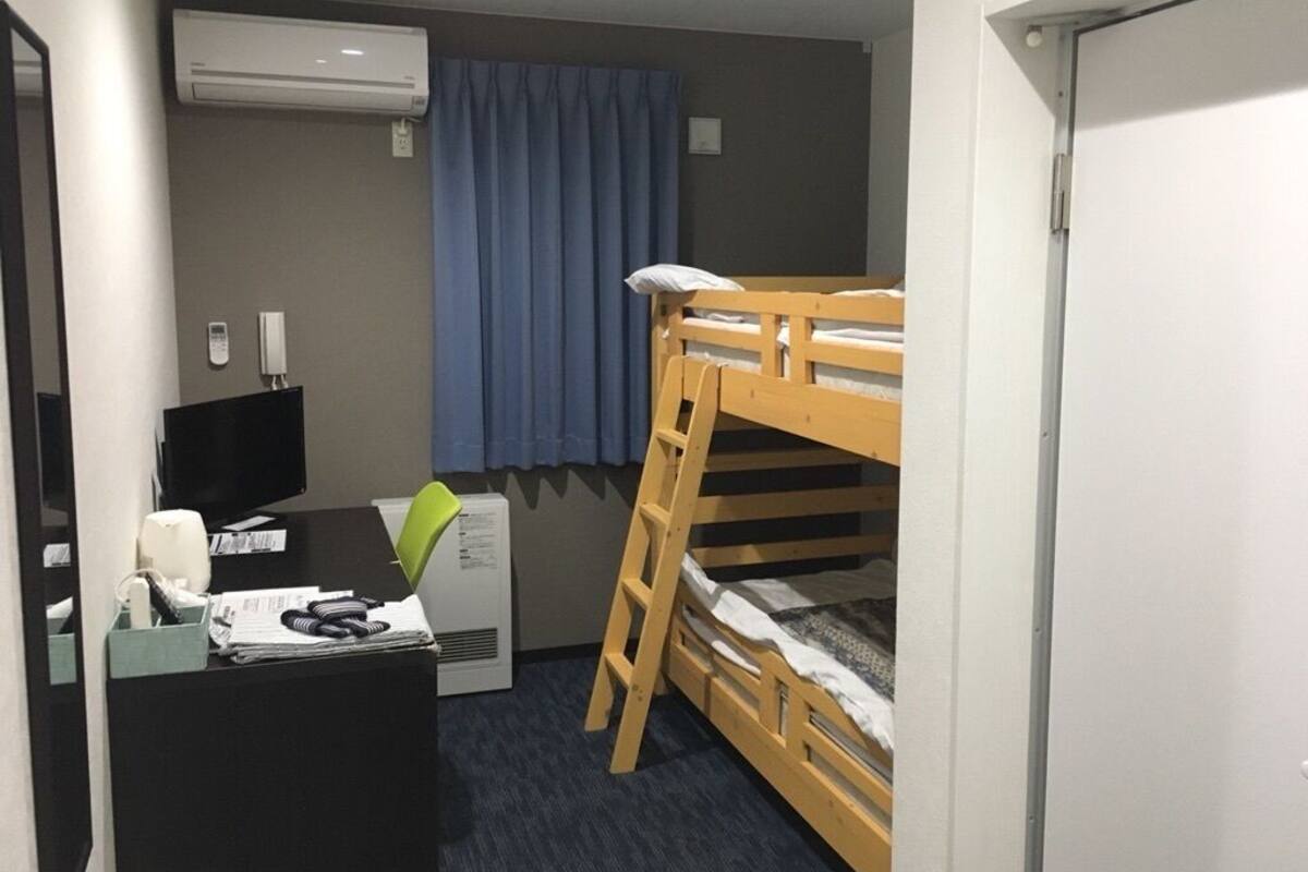 Habitación de hotel compacta con literas, un escritorio con silla, una lámpara de escritorio, teléfono y aire acondicionado.
