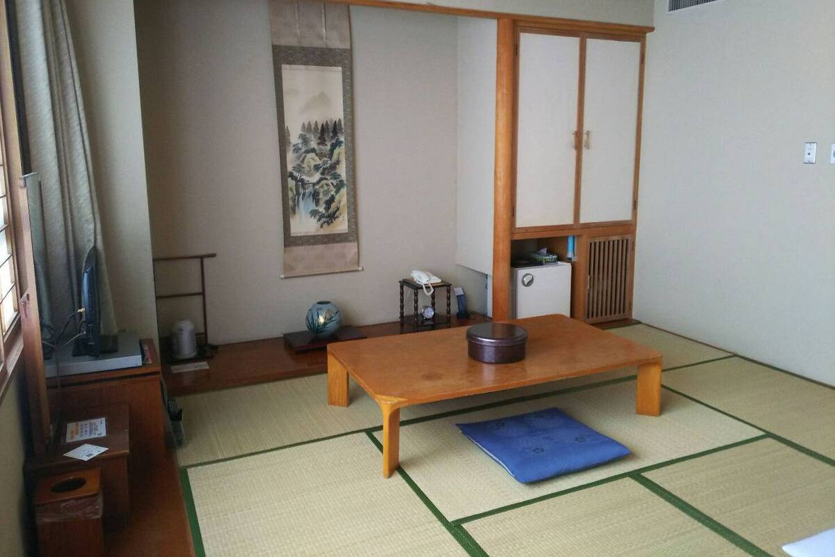 Una habitación japonesa tradicional con suelo de tatami, una mesa de madera baja, un tapete azul, un biombó shoji, un cuadro colgado y un pequeño armario.