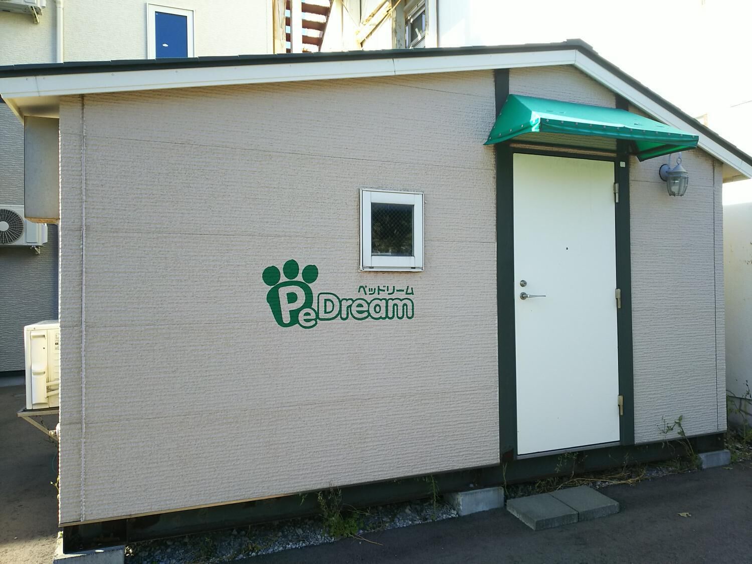 A small, single-story building with a beige exterior, a white door, and a small window. The building has a green awning and a logo with a paw print and the text "Pe Dream".