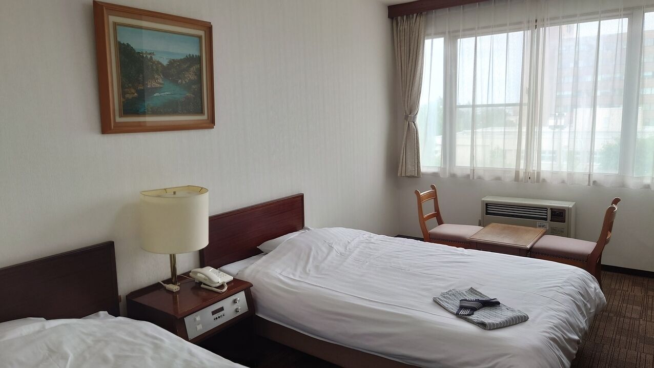 A hotel room with a bed, bedside table with a lamp and phone, a chair, a window with curtains, and an air conditioning unit.
