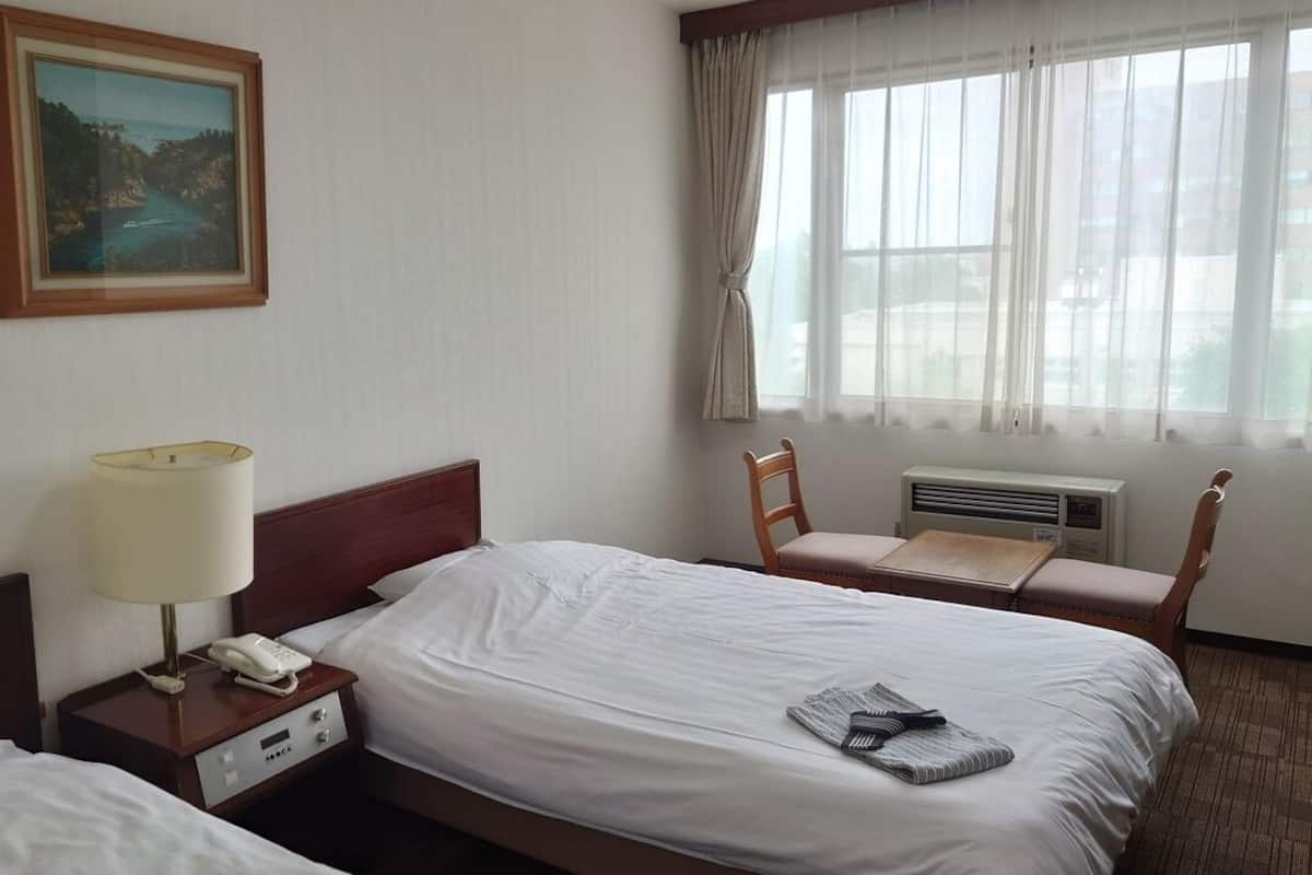 A hotel room with a bed, bedside table with a lamp and phone, a chair, a window with curtains, and an air conditioning unit.