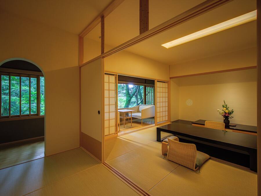 Een traditionele Japanse kamer met tatami vloeren, een lage tafel, een bank en een schuifdeur die uitkomt op een balkon met uitzicht op groen.
